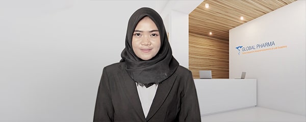 Seorang wanita berjilbab berdiri menghadap kamera sambil tersenyum, mengenakan blazer hitam; di belakangnya meja resepsionis kantor berlogo "GLOBAL PHARMA", dinding kayu dan pencahayaan hangat.