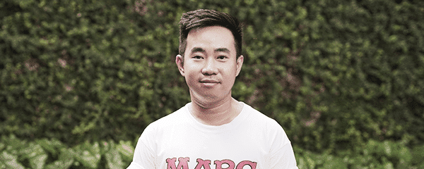 Seorang pria berdiri menghadap kamera dengan ekspresi netral, mengenakan kaus putih bertuliskan "MARC", berlatar hijau tanaman dan semak-semak di area luar ruangan.