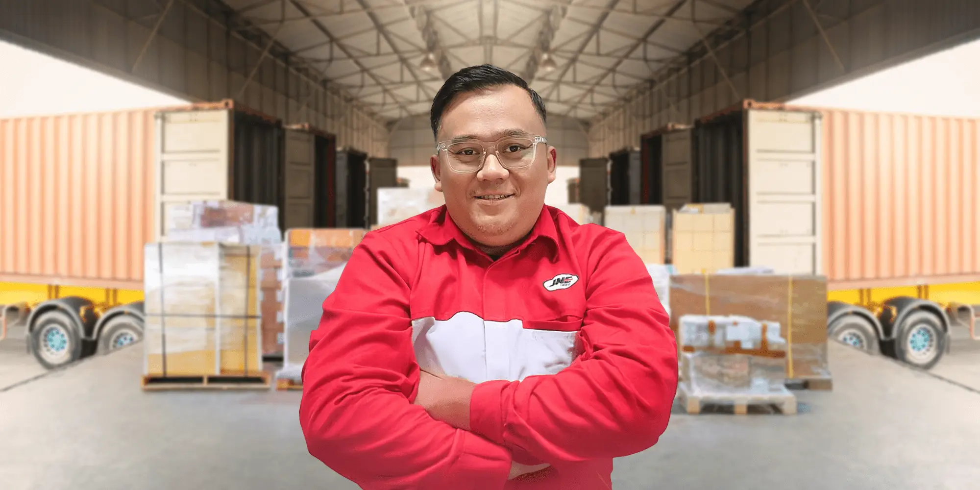 Seorang pria berseragam merah tersenyum sambil melipat lengan, berdiri di depan palet berisi kardus dan kontainer truk di dalam gudang penyimpanan.